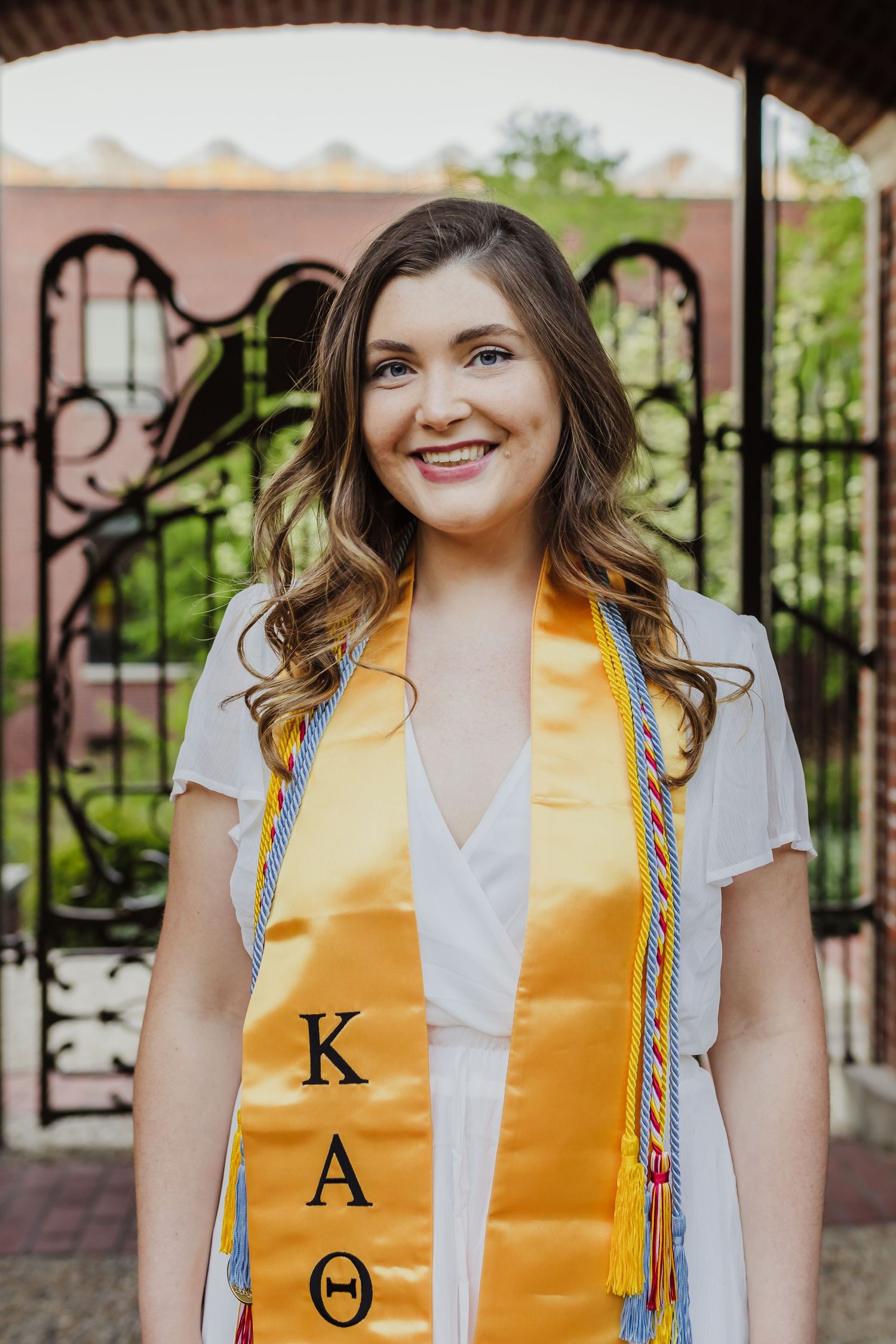 Graduate wearing gold stole with Greek letters poses under decorative arch during commencement.