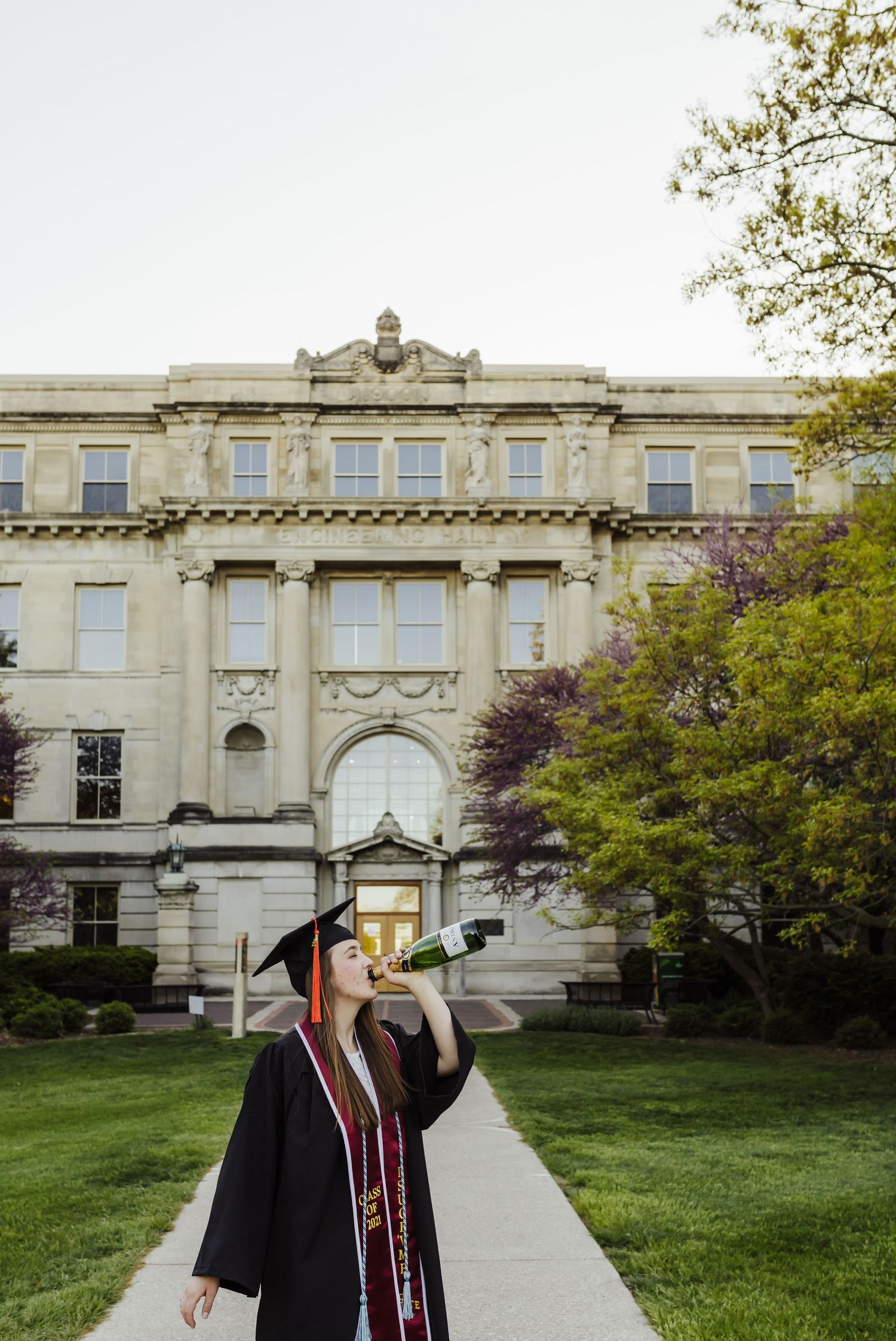 Graduate in cap and gown poses in front of historic university building with ornate architecture.