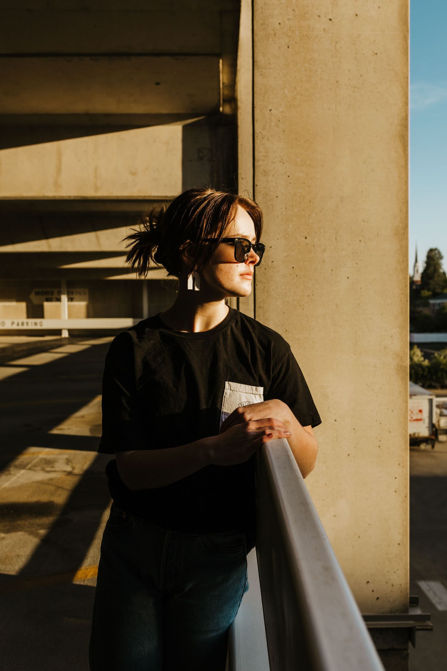 Portrait shot in golden sunlight against concrete columns wearing sunglasses and dark clothing.