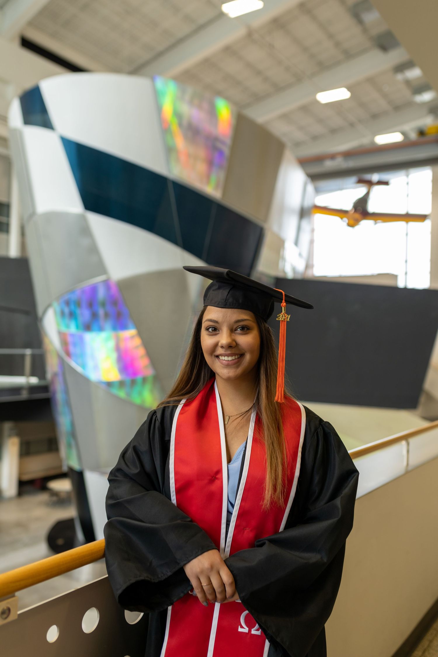 Graduate in red stole poses in modern building interior with holographic decor.