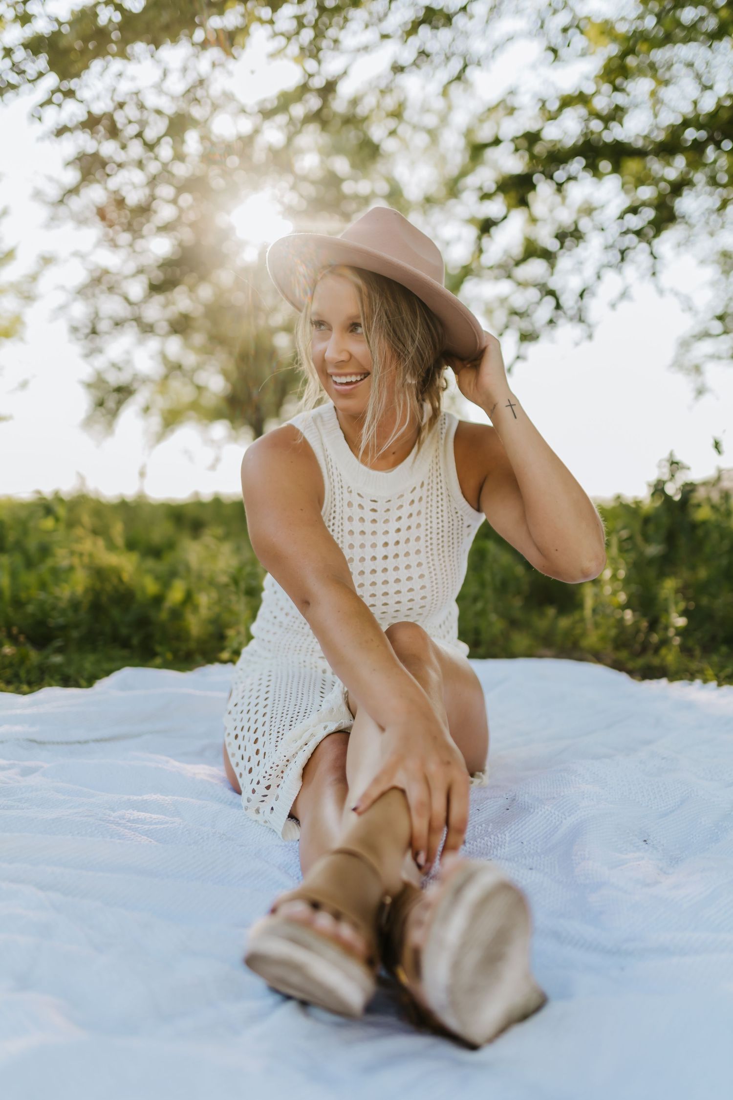 Woman in white crochet dress and hat sitting on blanket in sunlit field.