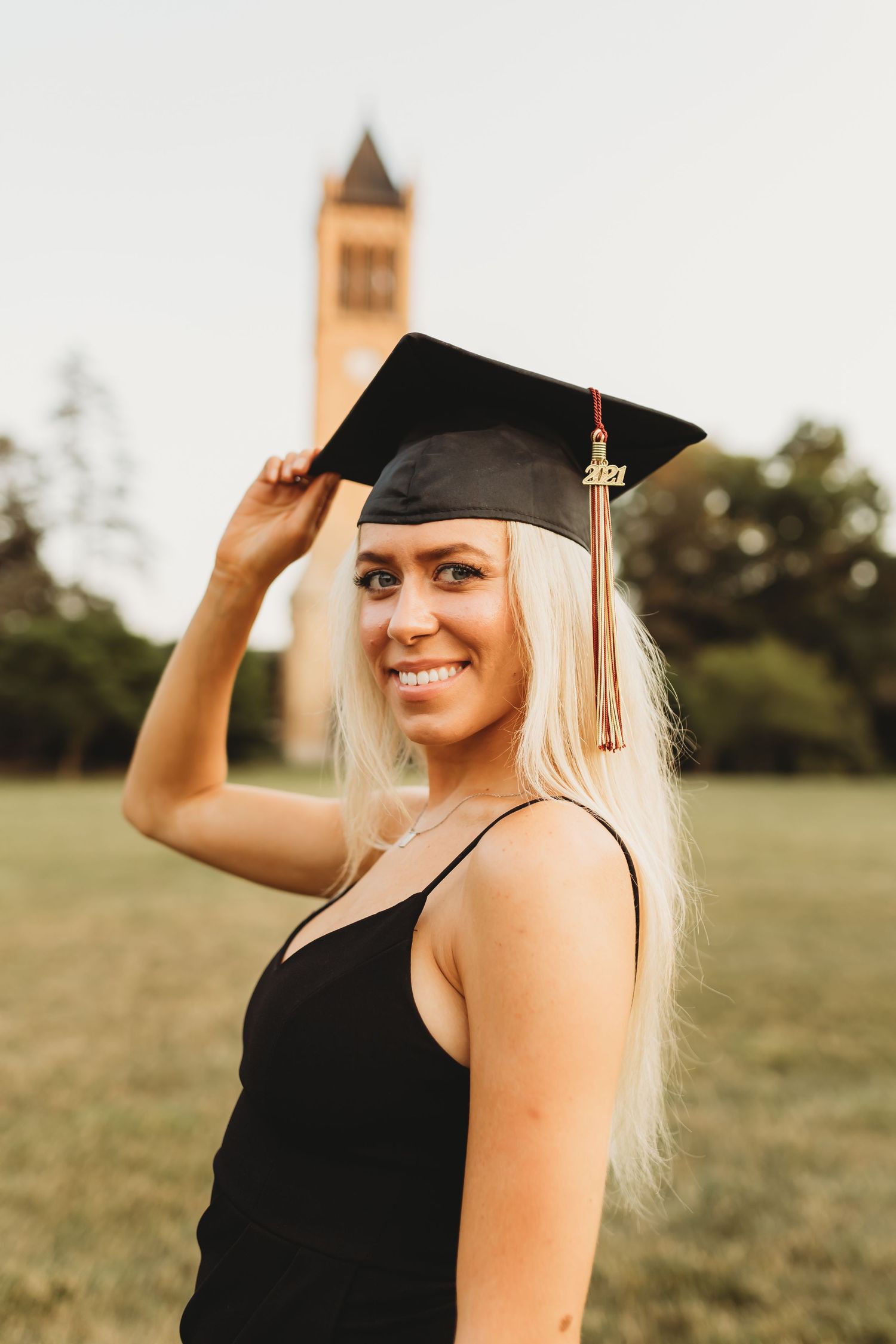 Graduate in black cap and gown poses in front of campus tower at sunset.