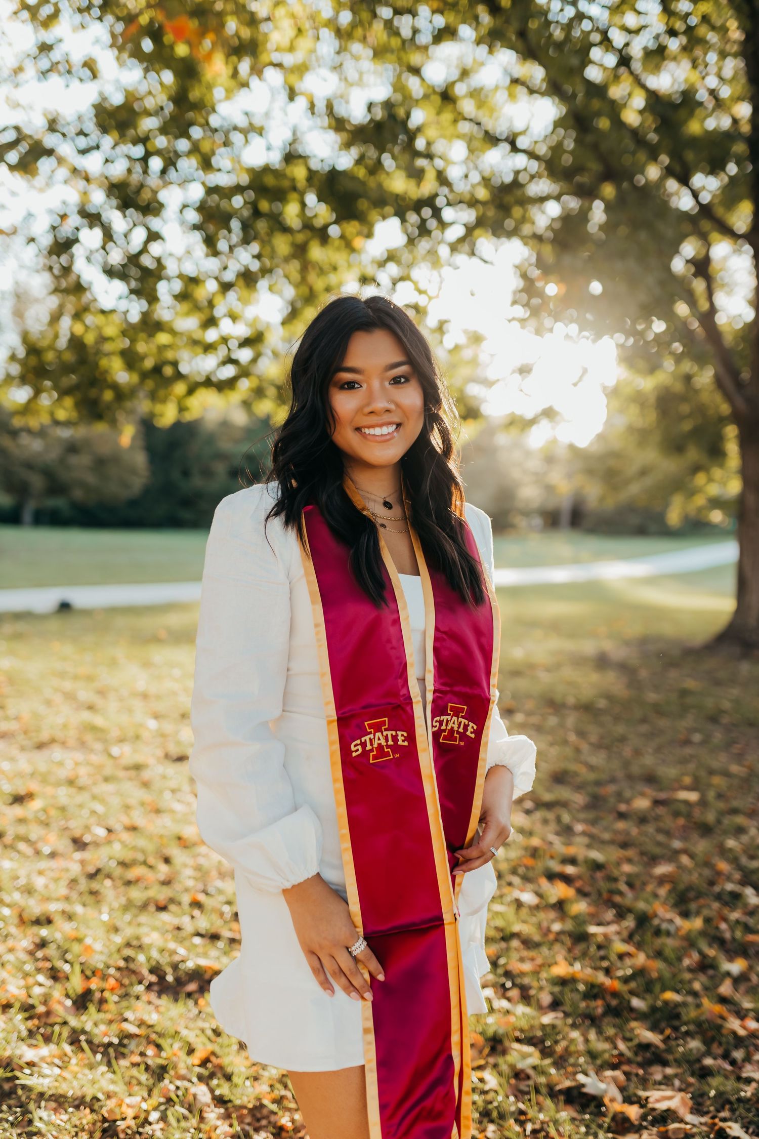 Graduate in white dress and red graduation stole poses outdoors during golden hour.