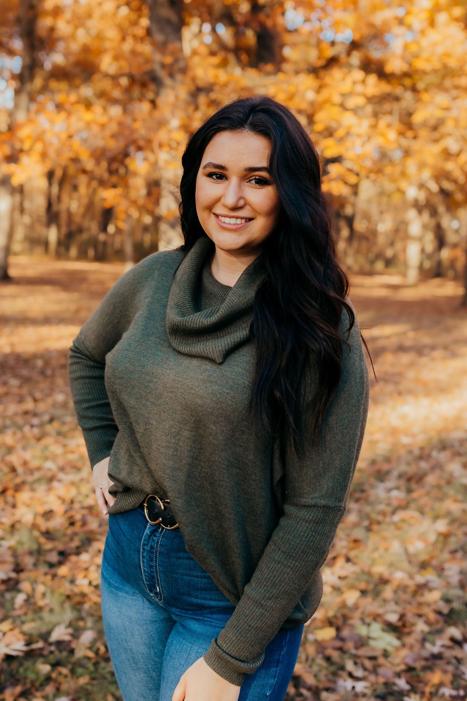 Woman in olive sweater smiles while standing amid fall foliage.