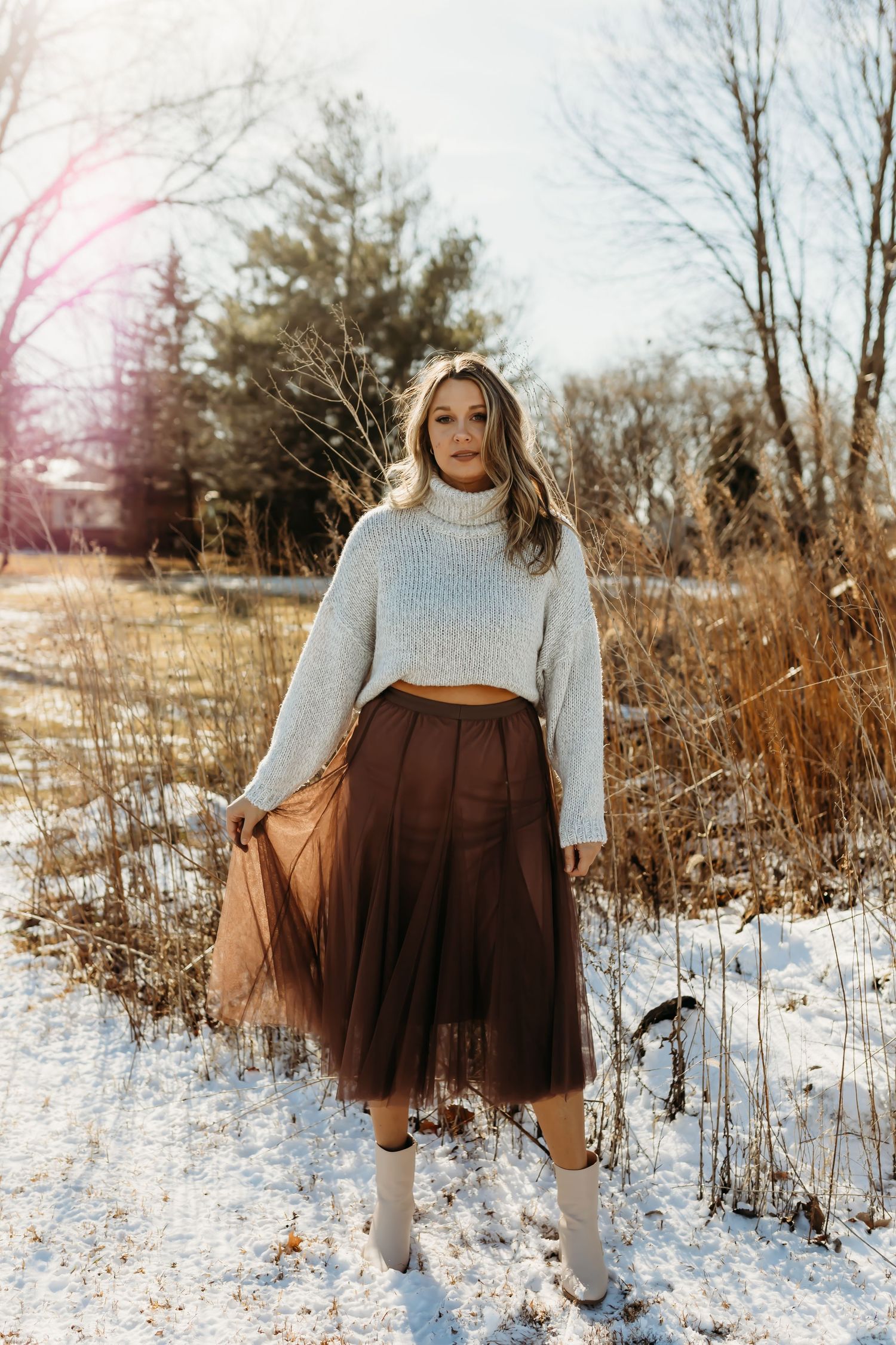 Winter fashion portrait of woman in white sweater and brown skirt standing in snowy field.