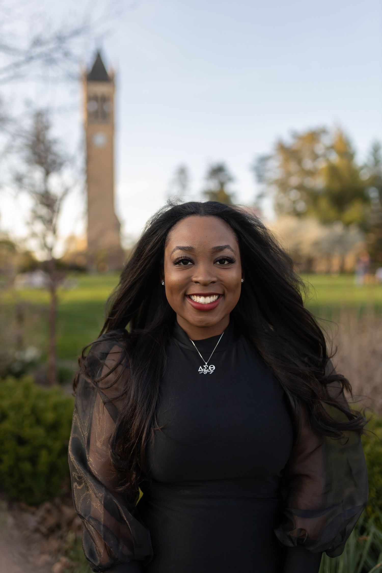 Graduate in elegant black dress poses in front of iconic campus tower during golden hour.