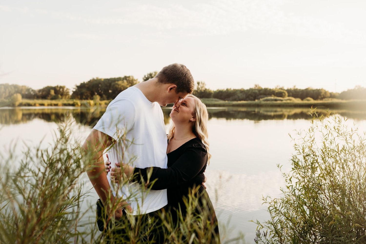 A romantic couple embracing by a peaceful lake at sunset surrounded by tall grass.