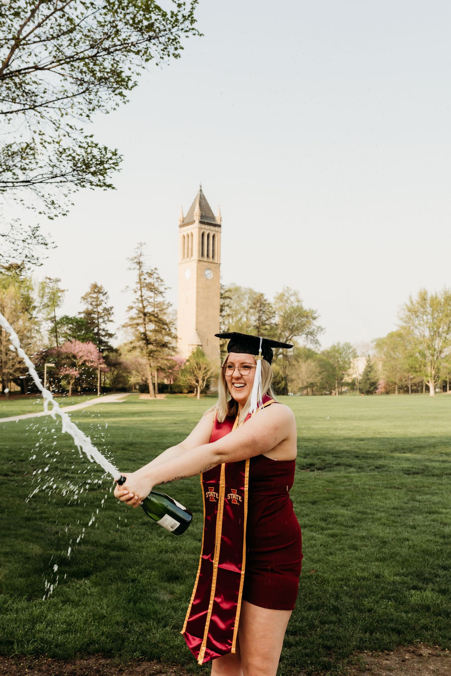 Graduate in red dress popping champagne bottle on campus lawn with clock tower in background.