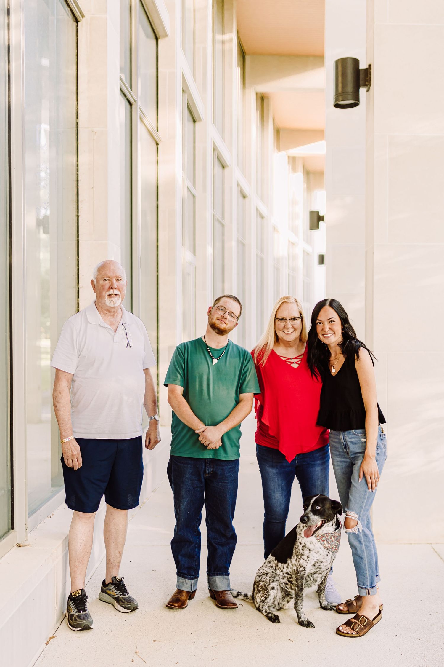 A family group poses casually with their spotted dog along a modern building exterior.