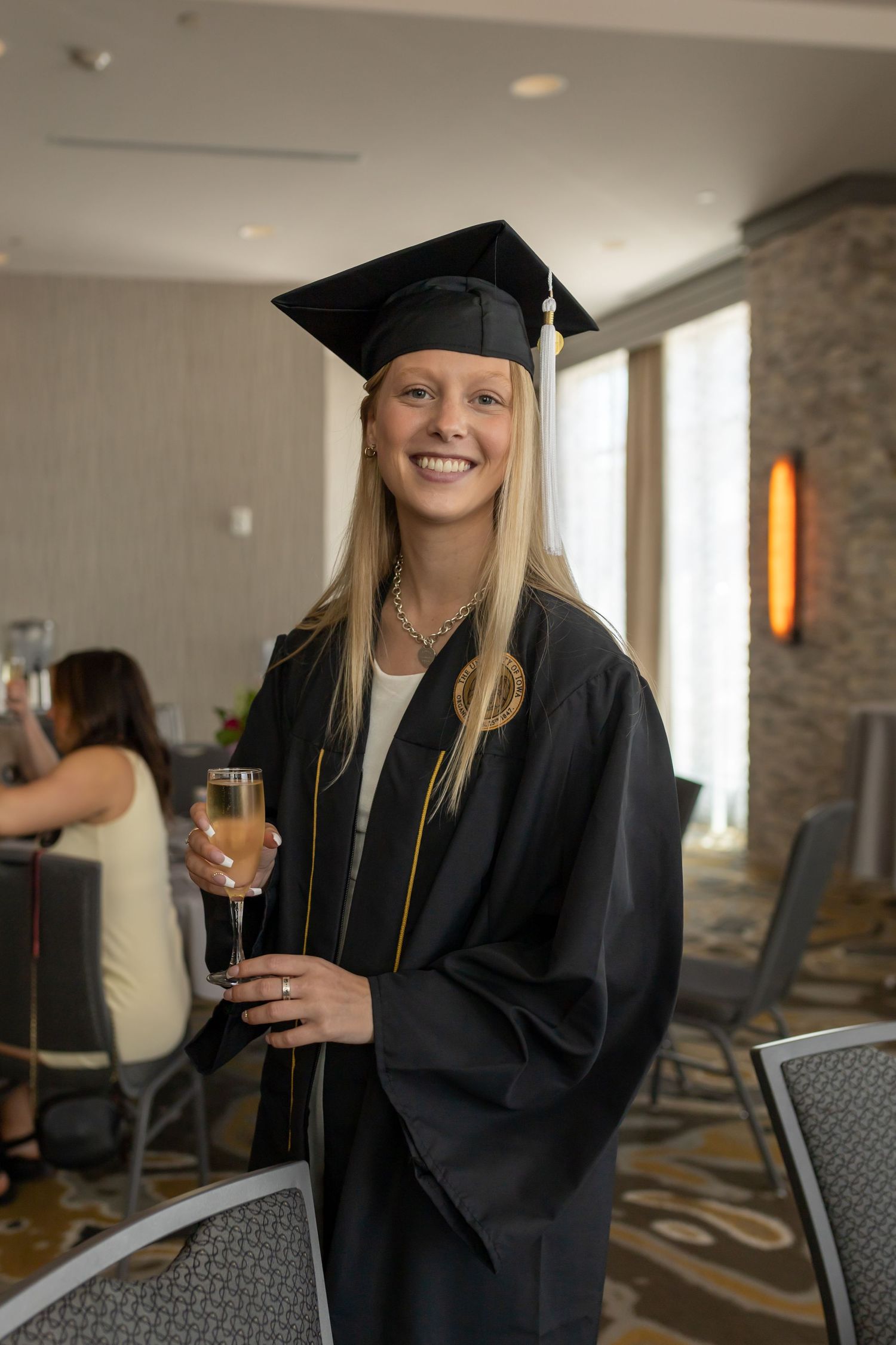 Graduate in traditional cap and gown holds champagne glass at indoor celebration.