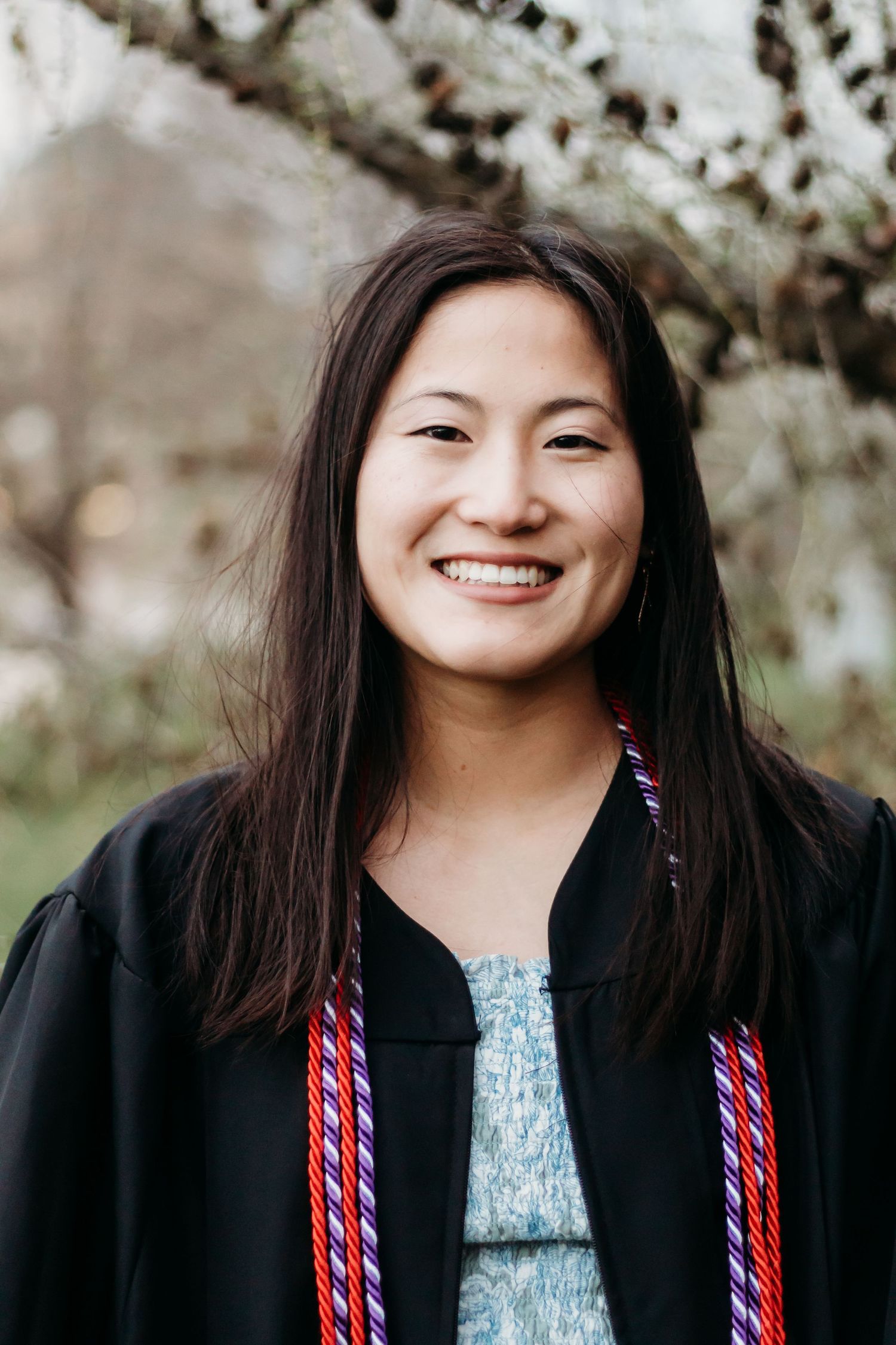 Graduate in traditional black gown with colorful stole smiling against blooming spring background.