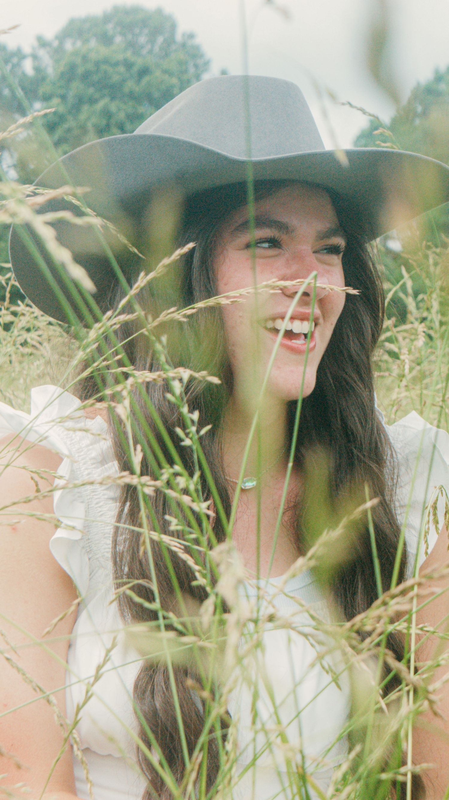 A person wearing a cowboy hat smiling in tall grass with soft natural lighting.