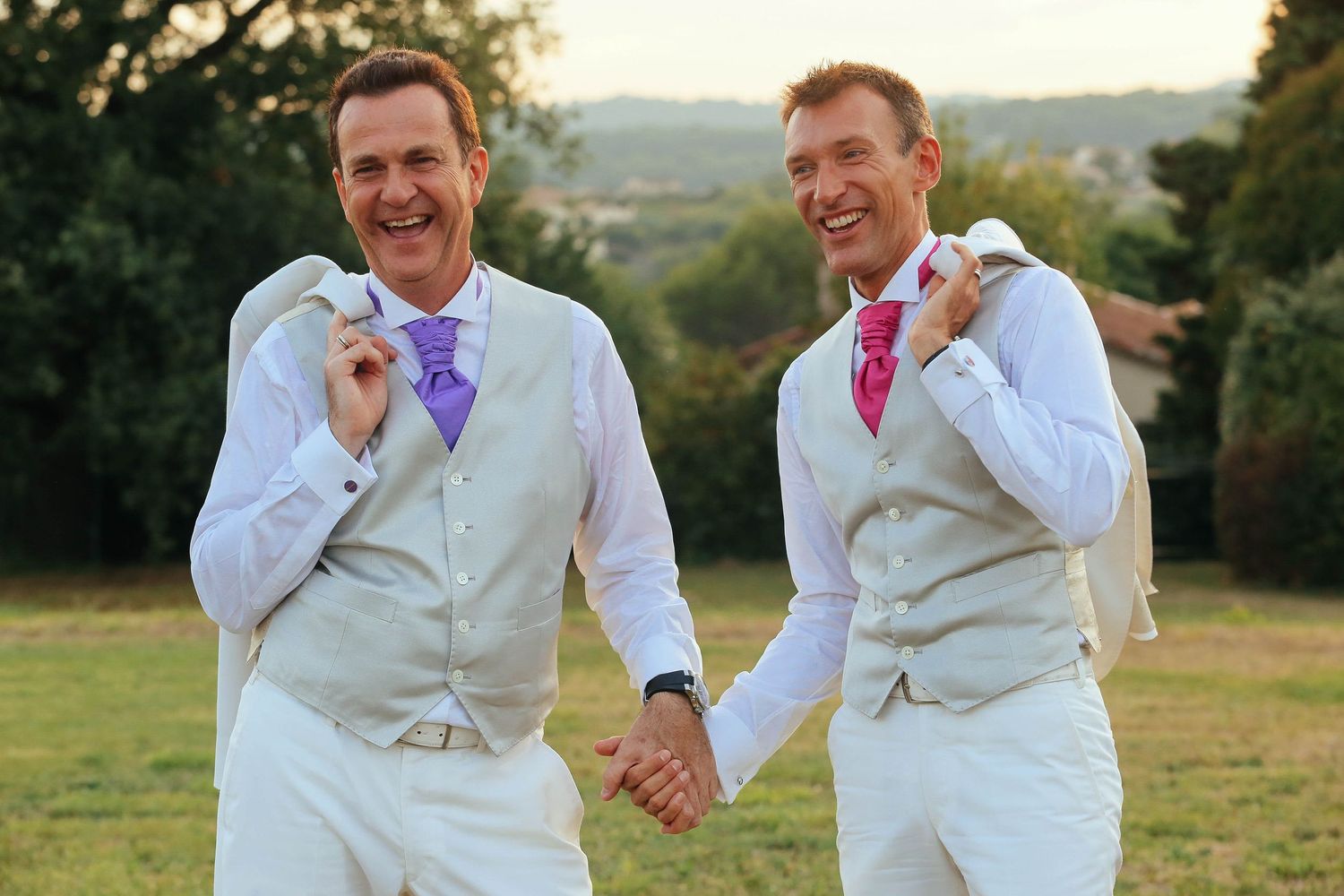 Two grooms in matching light-colored suits embracing outdoors on their wedding day.