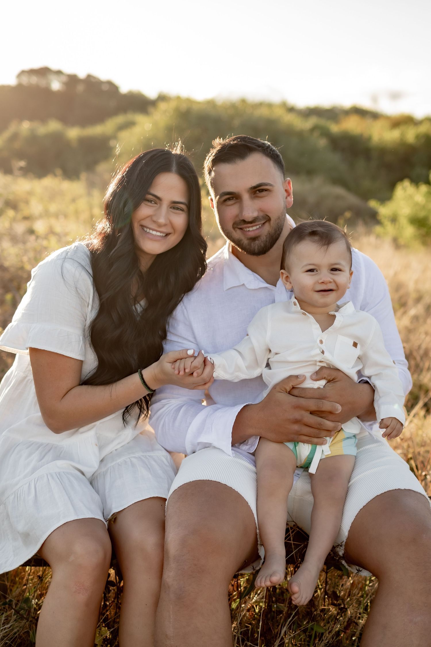 A happy family of three wearing white sits together on a rustic bench in golden sunlight.
