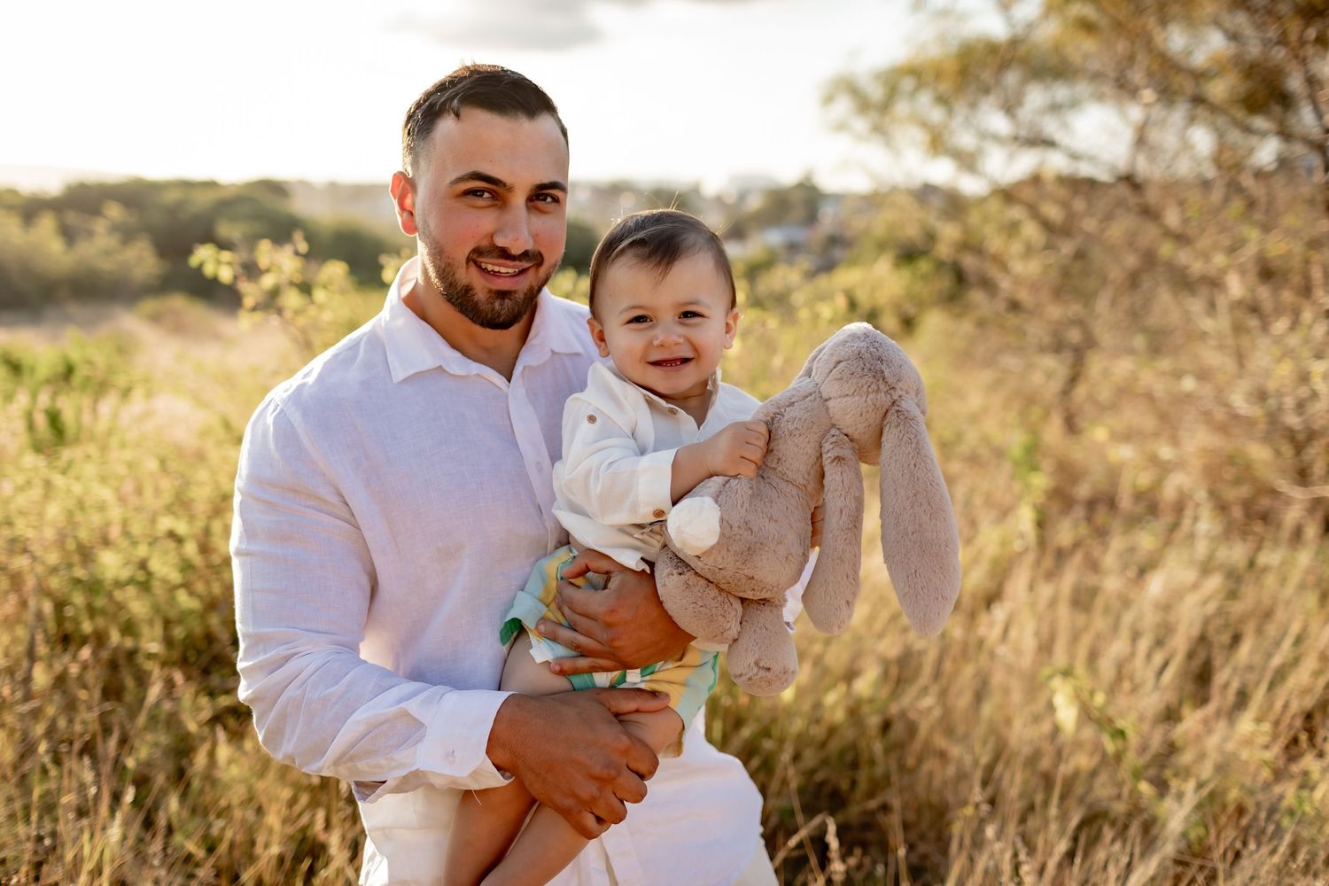 Parent and smiling baby holding stuffed toy outdoors in golden sunlight.