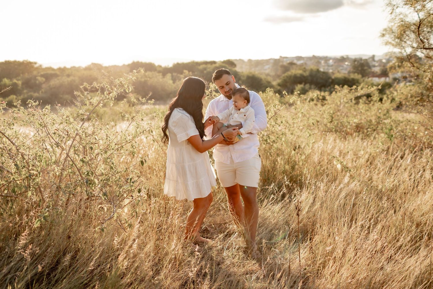 A couple shares a tender moment with their baby in a sun-drenched field of tall grass.