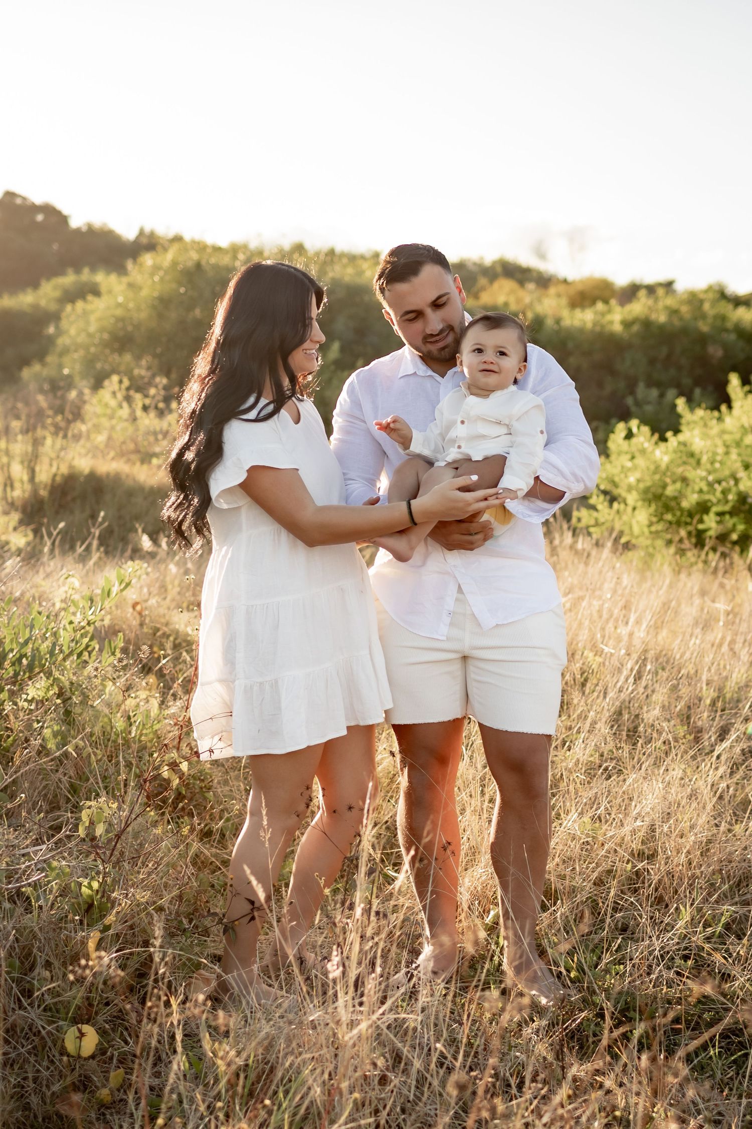 A family in matching white outfits stands together in a sun-lit field of tall grass.