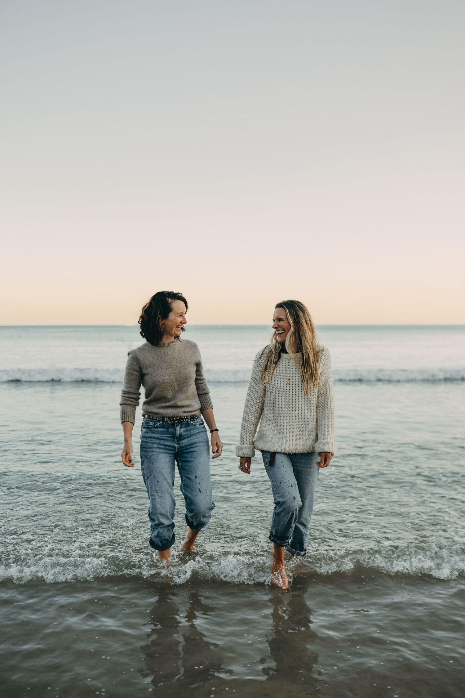 Two friends walking barefoot in shallow ocean waves at sunset.