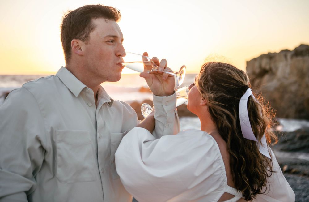 A couple shares drinks together against a sunset backdrop by the ocean.