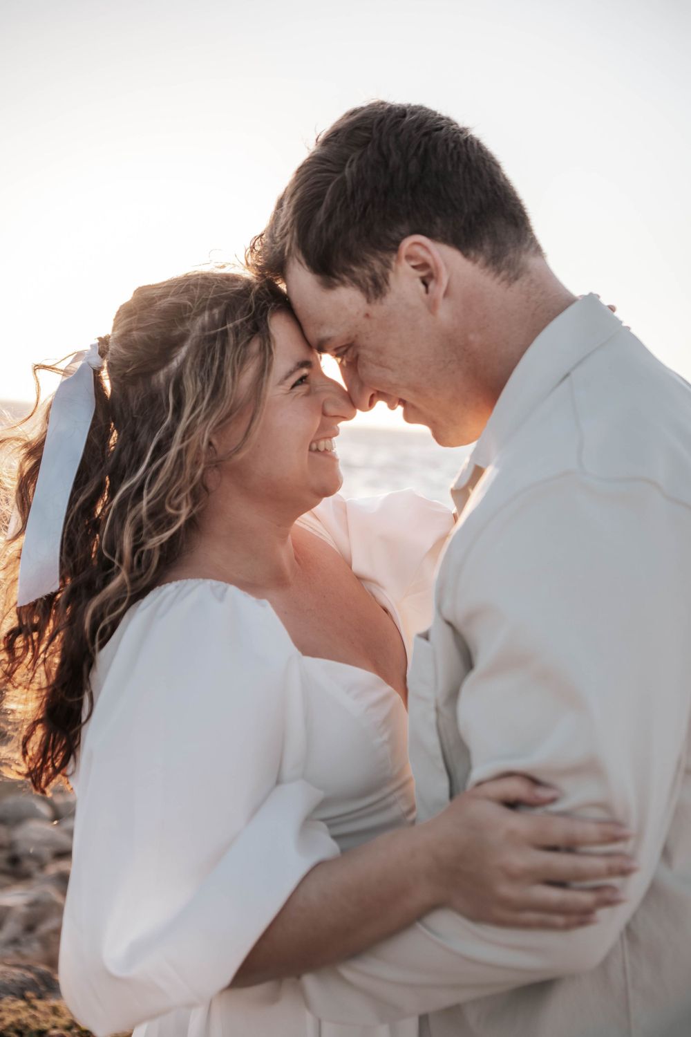 A couple in white outfits embraces tenderly during golden hour.