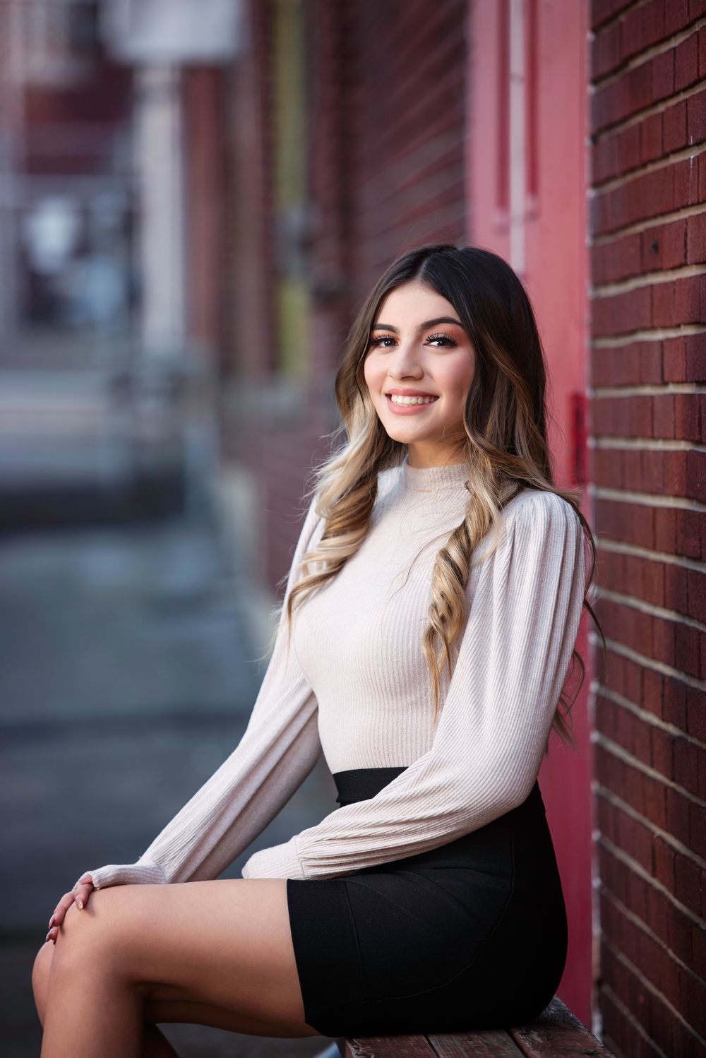 Brunette teen in cream ribbed turtleneck and black skirt sits smiling against brick wall.