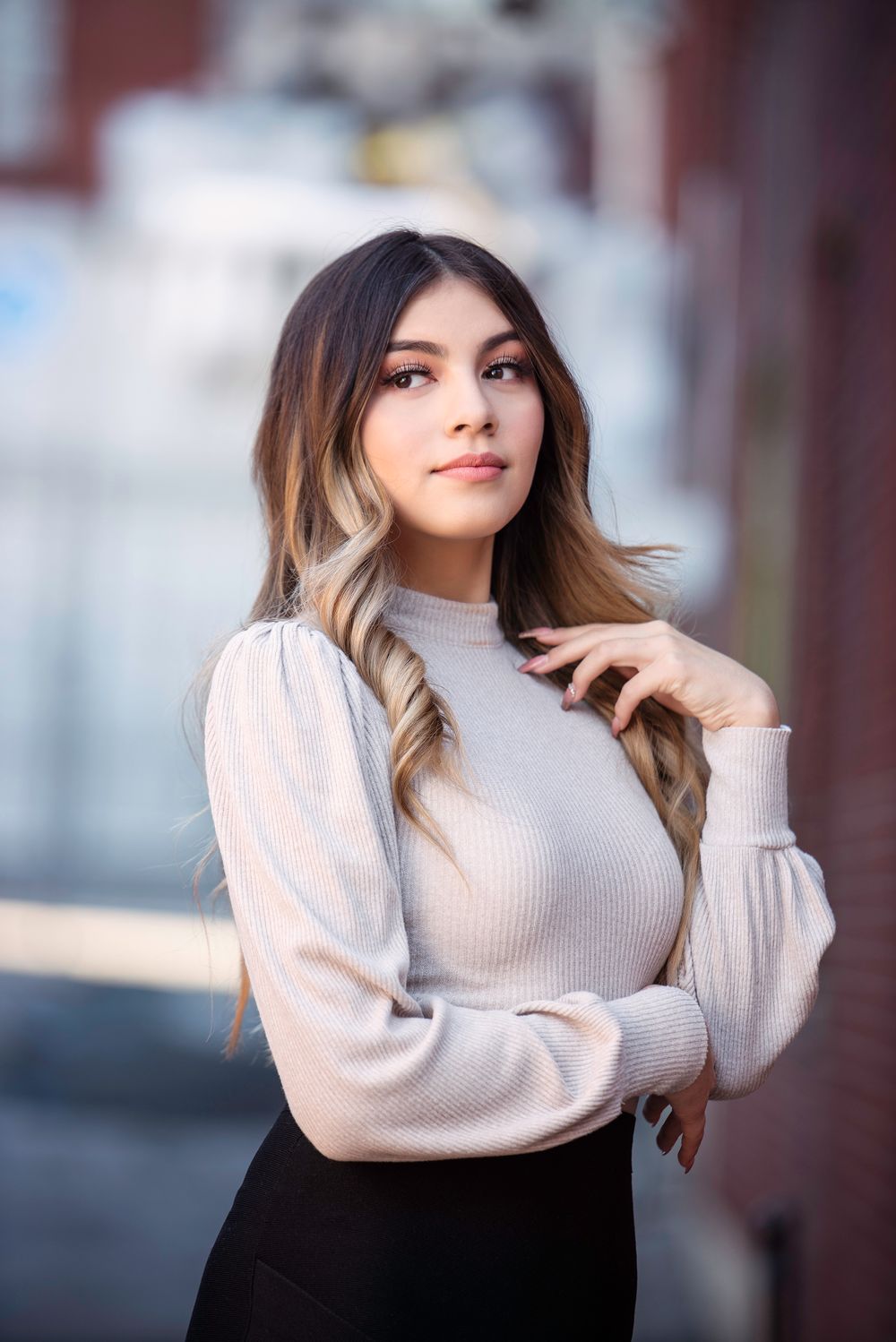 Young woman in cream turtleneck sweater with ombre hair posing against blurred brick background.