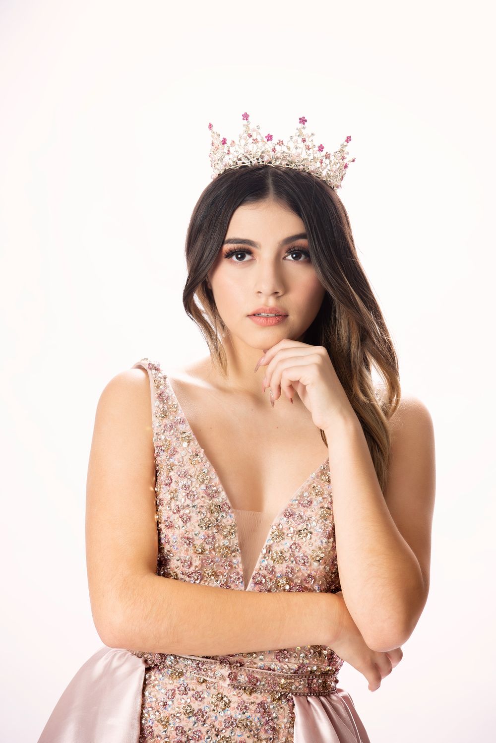 Young woman wearing crystal crown and pink floral gown posing thoughtfully against white background.