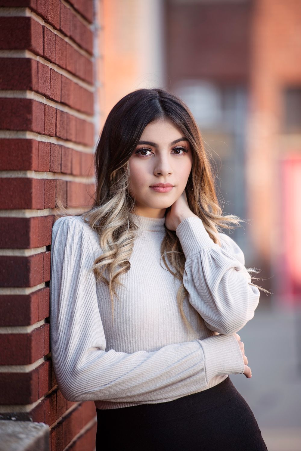 Dark-haired teen in cream ribbed turtleneck leans against red brick wall in urban setting.