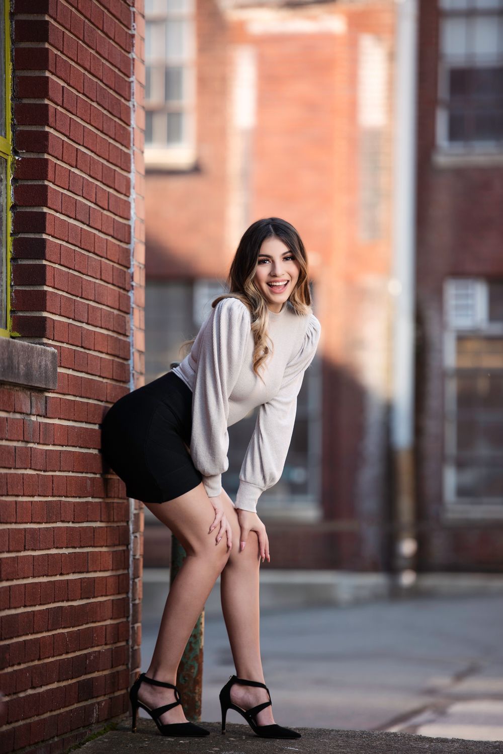 Young woman in white sweater and black mini skirt posing against brick wall in urban setting.