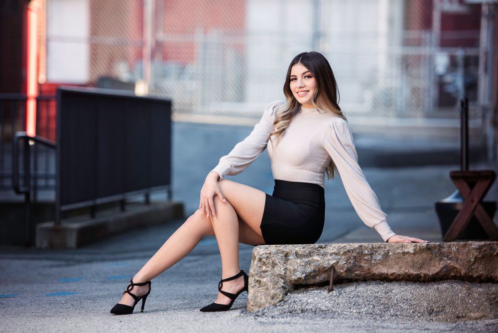Stylish young woman in beige top and black mini skirt sitting on stone ledge outdoors.