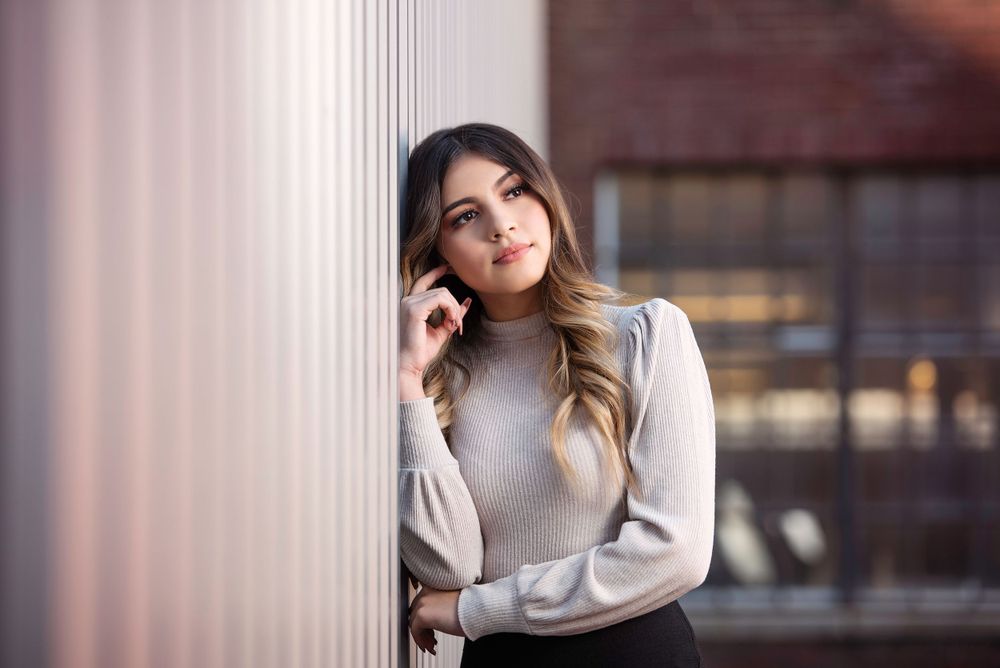 Elegant young woman in grey turtleneck sweater leaning against white column in urban setting.