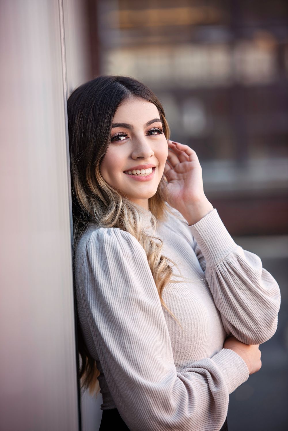 Smiling young woman in cream ribbed sweater leaning against white wall in close-up portrait.