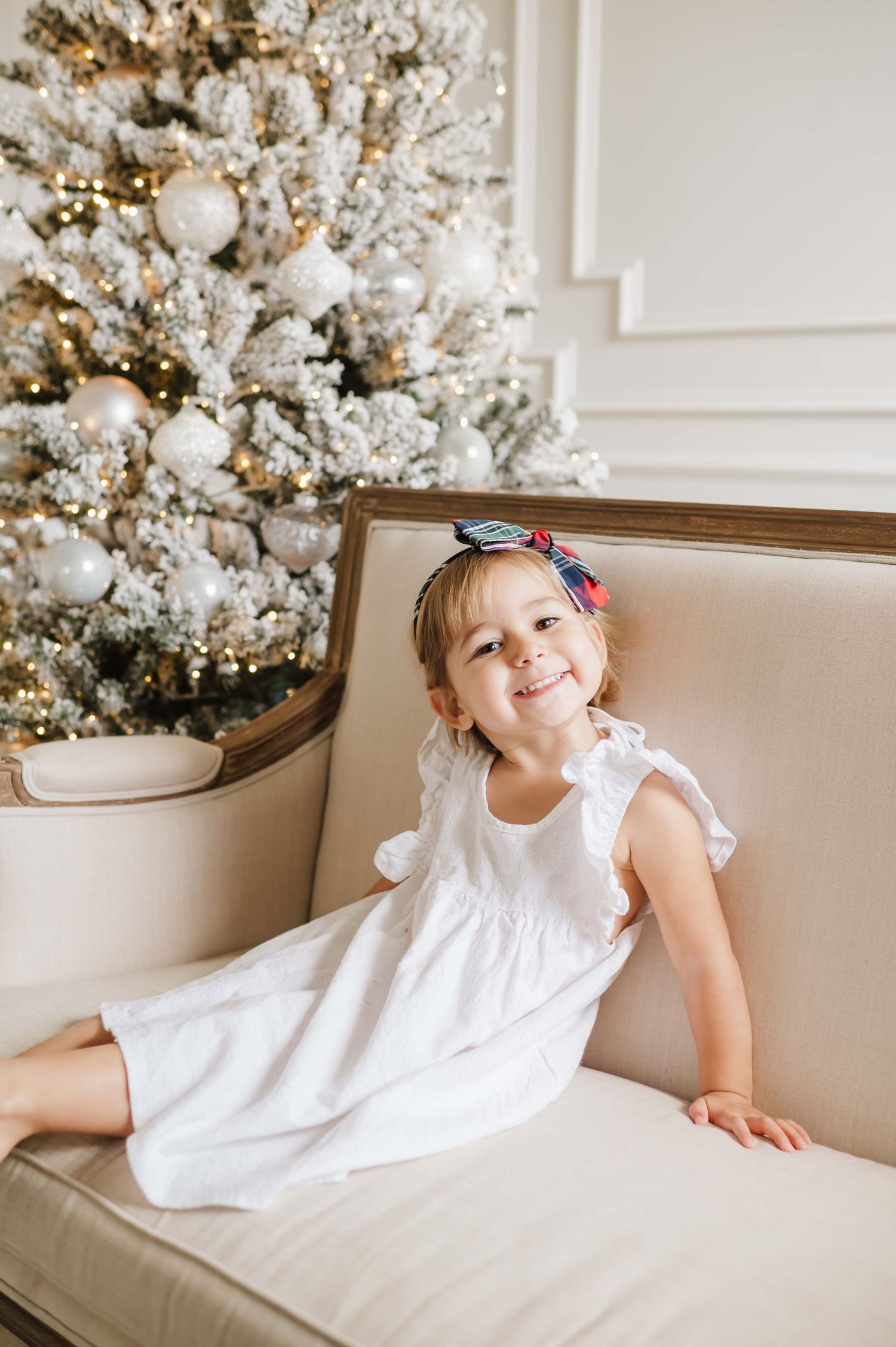 toddler posing for a christmas mini photoshoot in a studio with a plaid red headband and a white linen christmas pjs