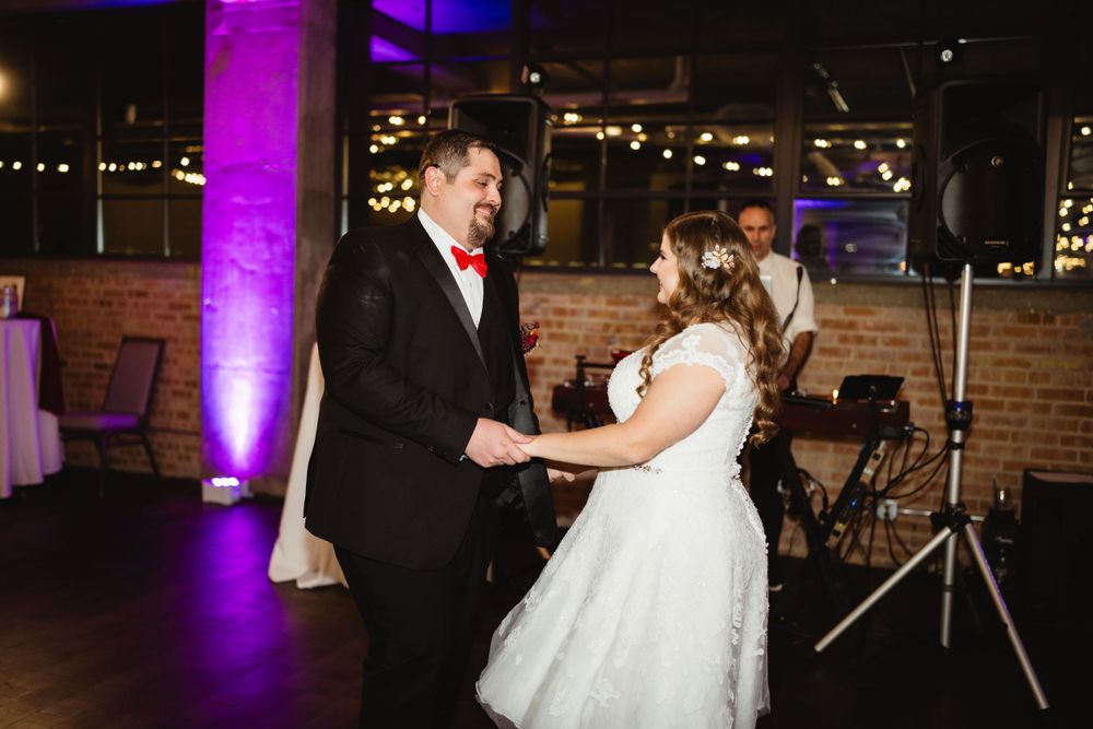 A couple shares their first dance at a wedding reception illuminated by purple uplighting in an industrial venue.