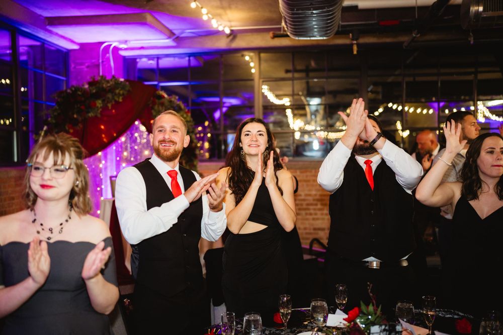 Wedding guests clapping and celebrating during a reception with purple mood lighting and industrial decor.