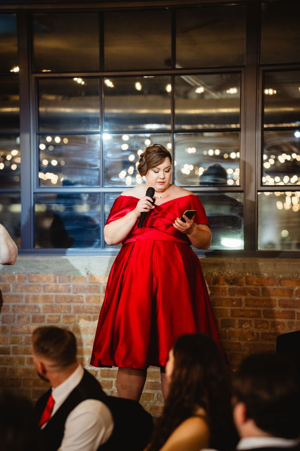 Woman in a red dress gives a speech at an event space with exposed brick walls and city lights in the background.