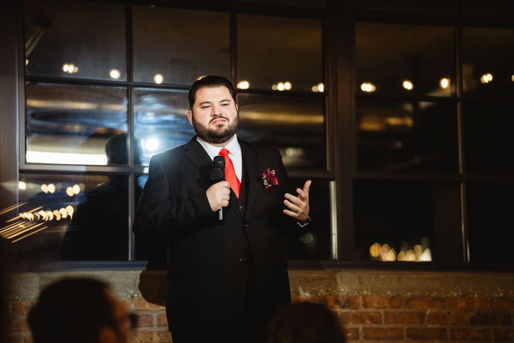Speaker in formal attire addresses guests at an evening event in an industrial-style venue.