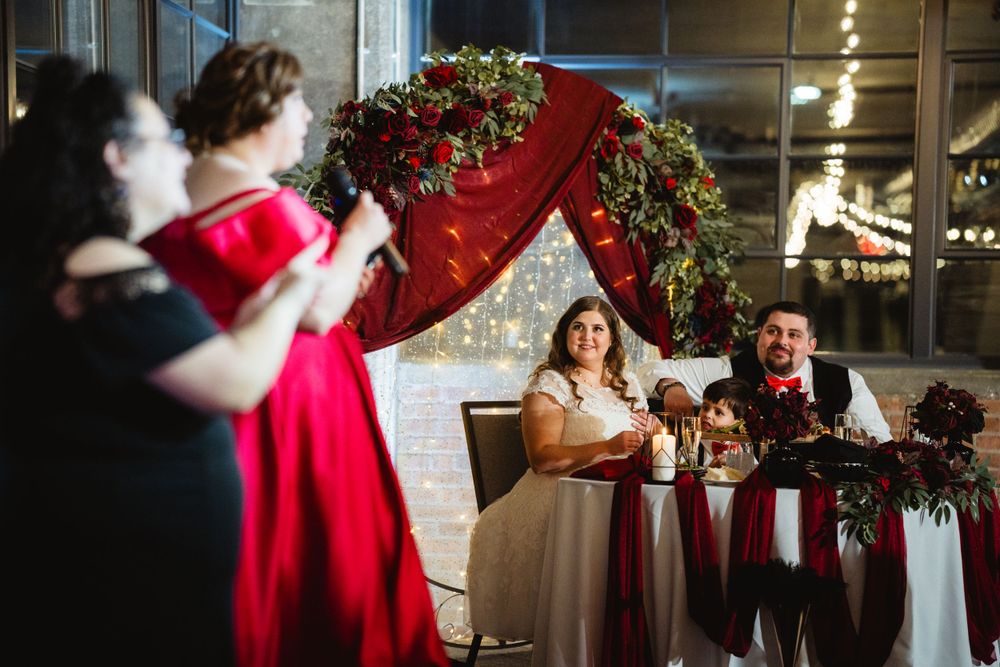 Wedding celebration under a red floral arch in an industrial-style venue with brick walls and large windows.