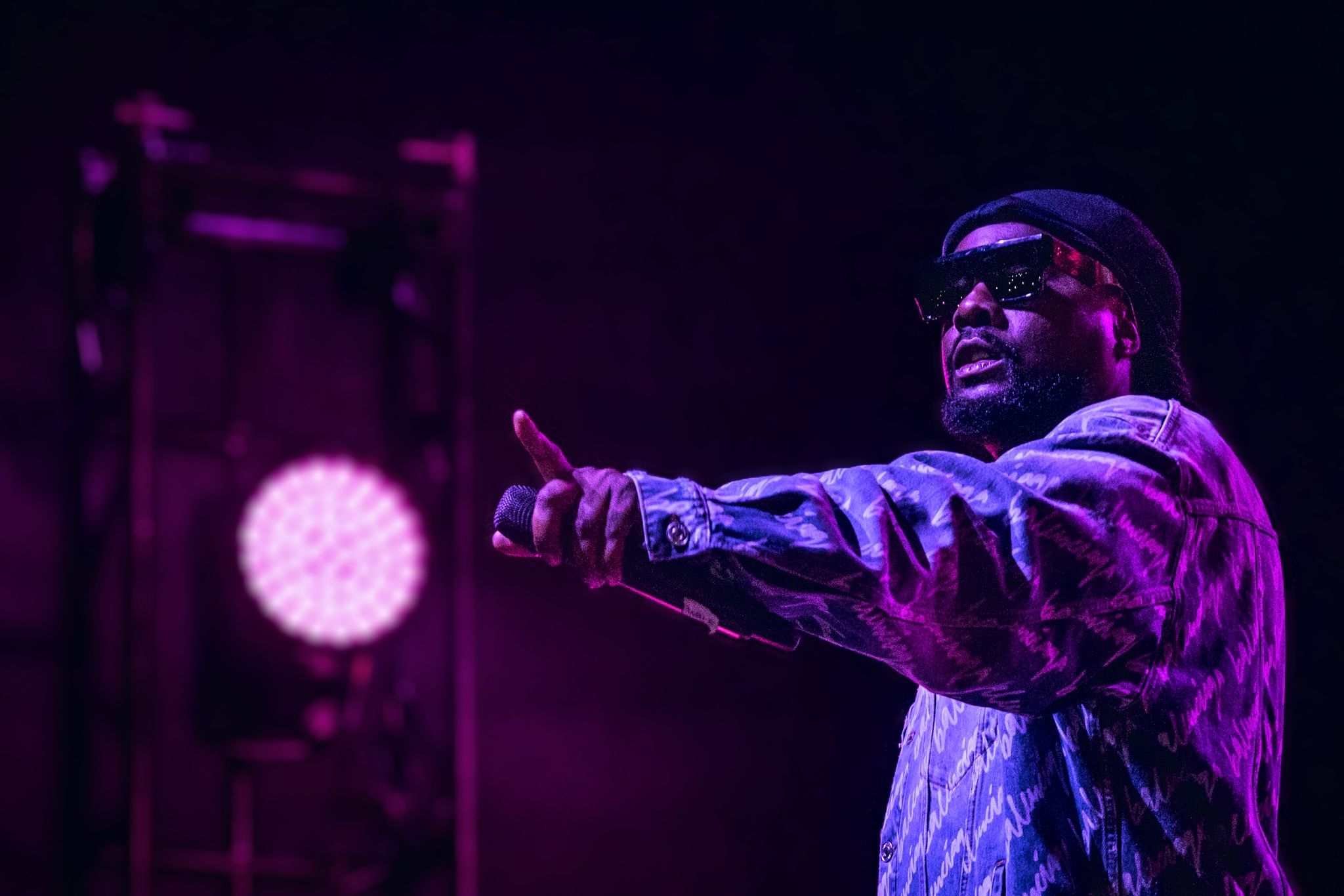 Concert performer in camouflage jacket pointing under purple stage lighting.