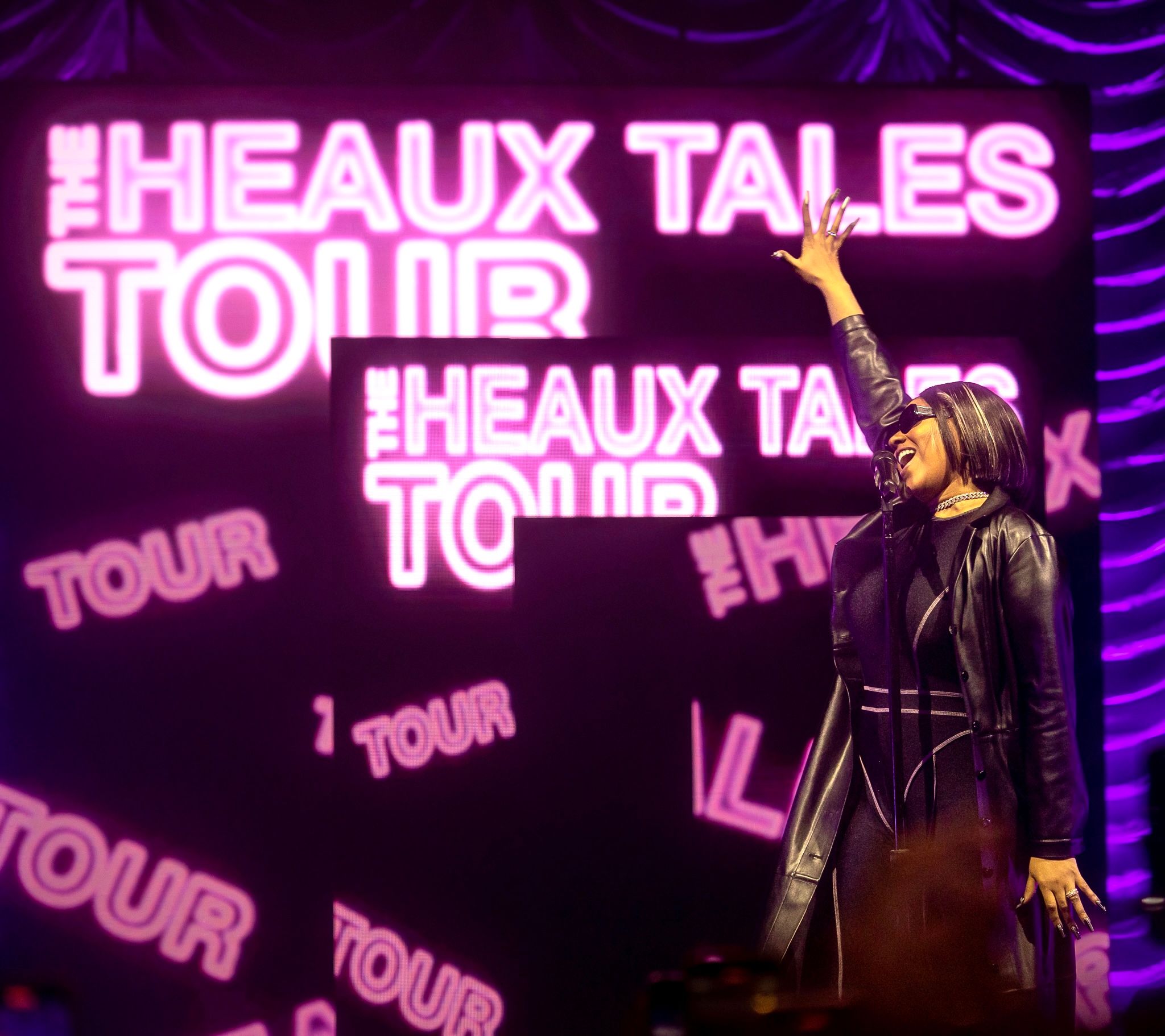 Performer silhouetted against pink neon 'Heaux Tales Tour' signage on stage.