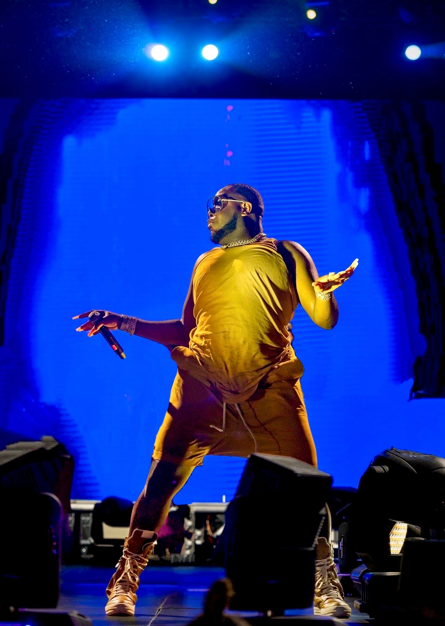 Performer in yellow outfit striking dramatic pose against blue-lit backdrop on concert stage.