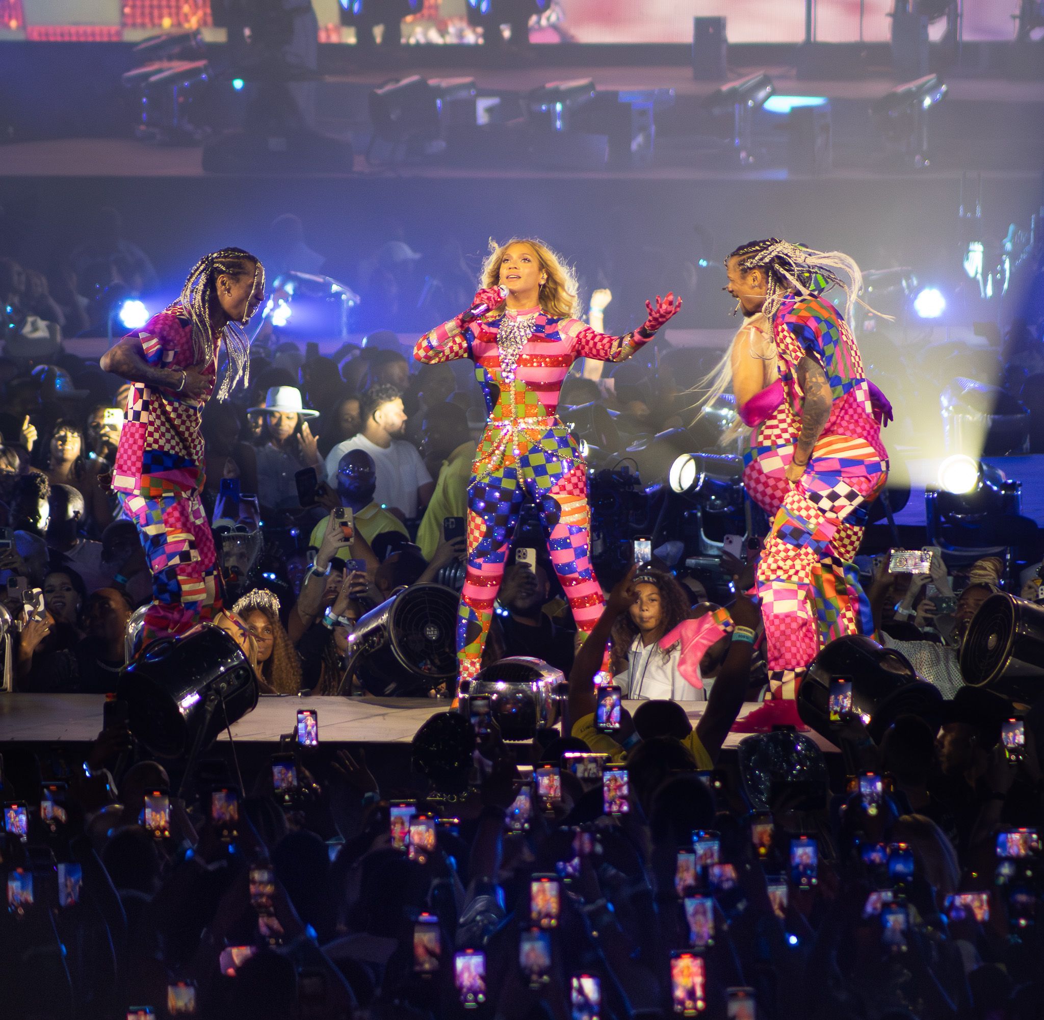 Dancers in colorful costumes perform energetic choreography on stage with audience phones lighting up below.