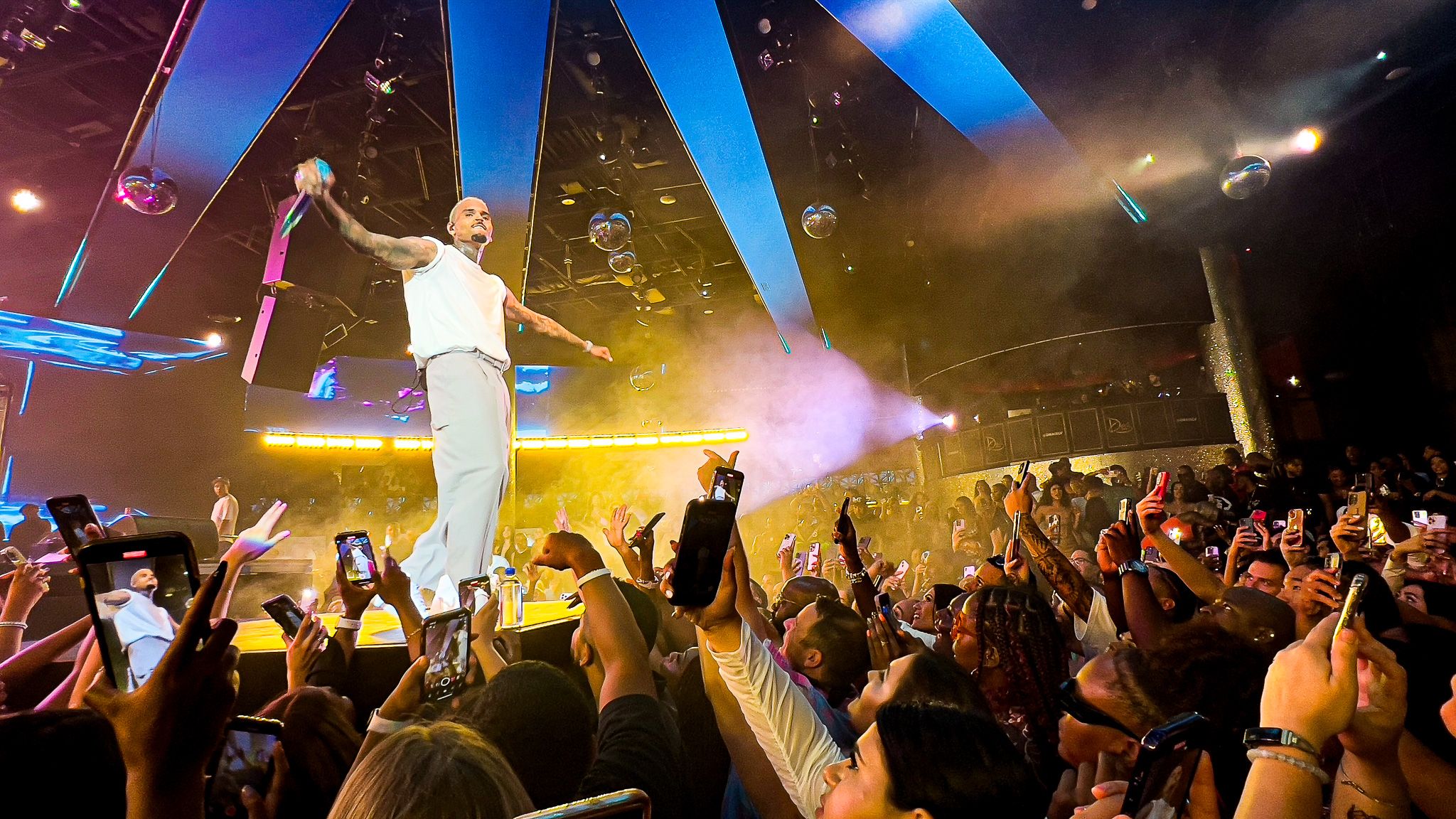 Concert performer in white outfit stands elevated above cheering crowd with dramatic blue lighting effects.