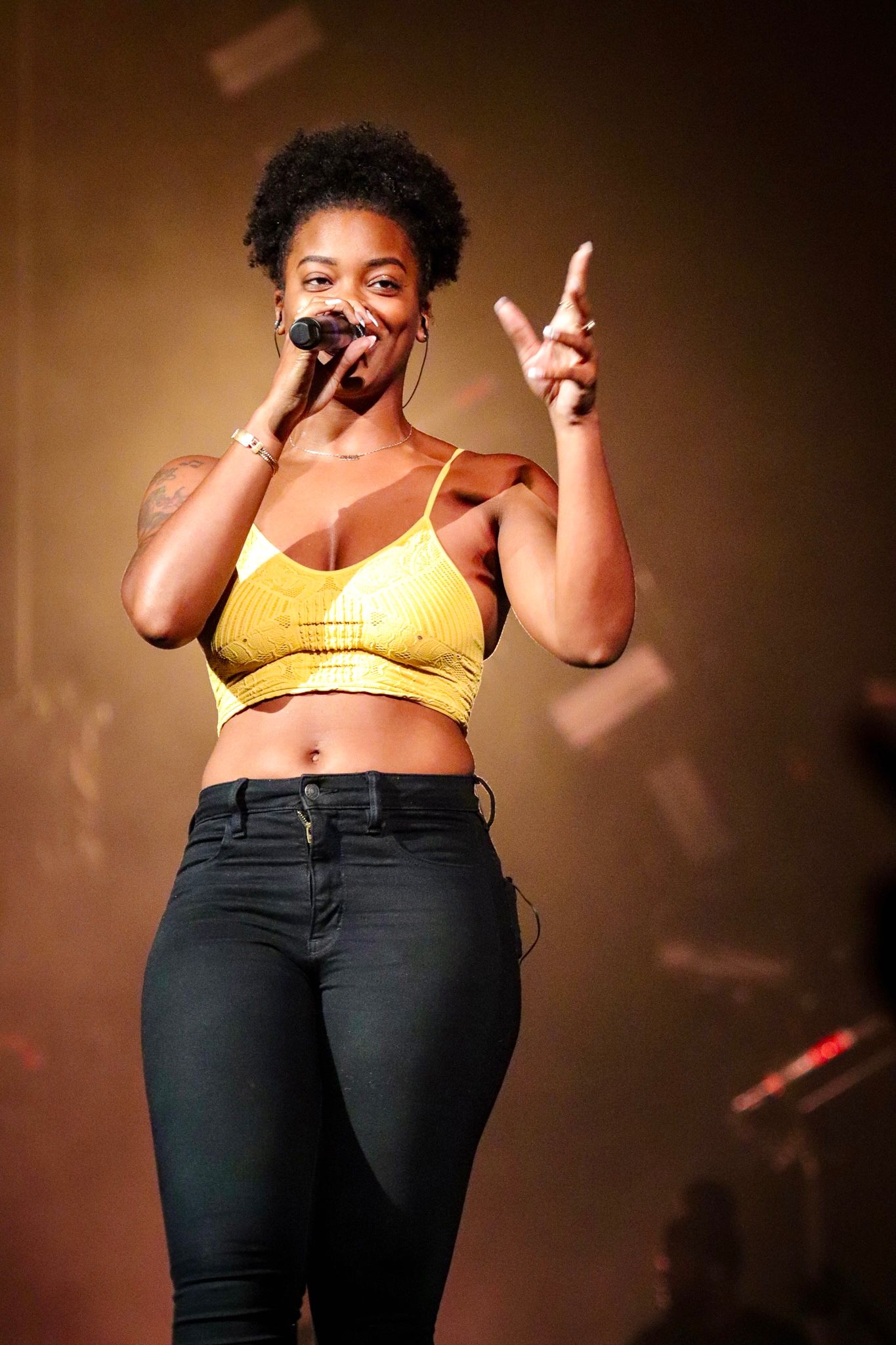 Concert performer in yellow crop top and black jeans gestures while singing on warm-lit stage.
