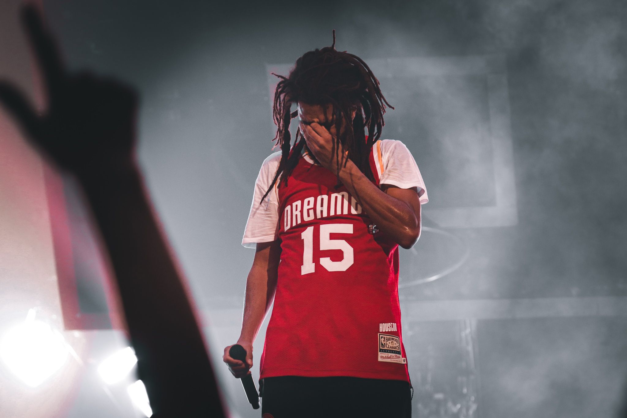 Concert performer in red number 15 jersey dramatically illuminated on smoky stage.