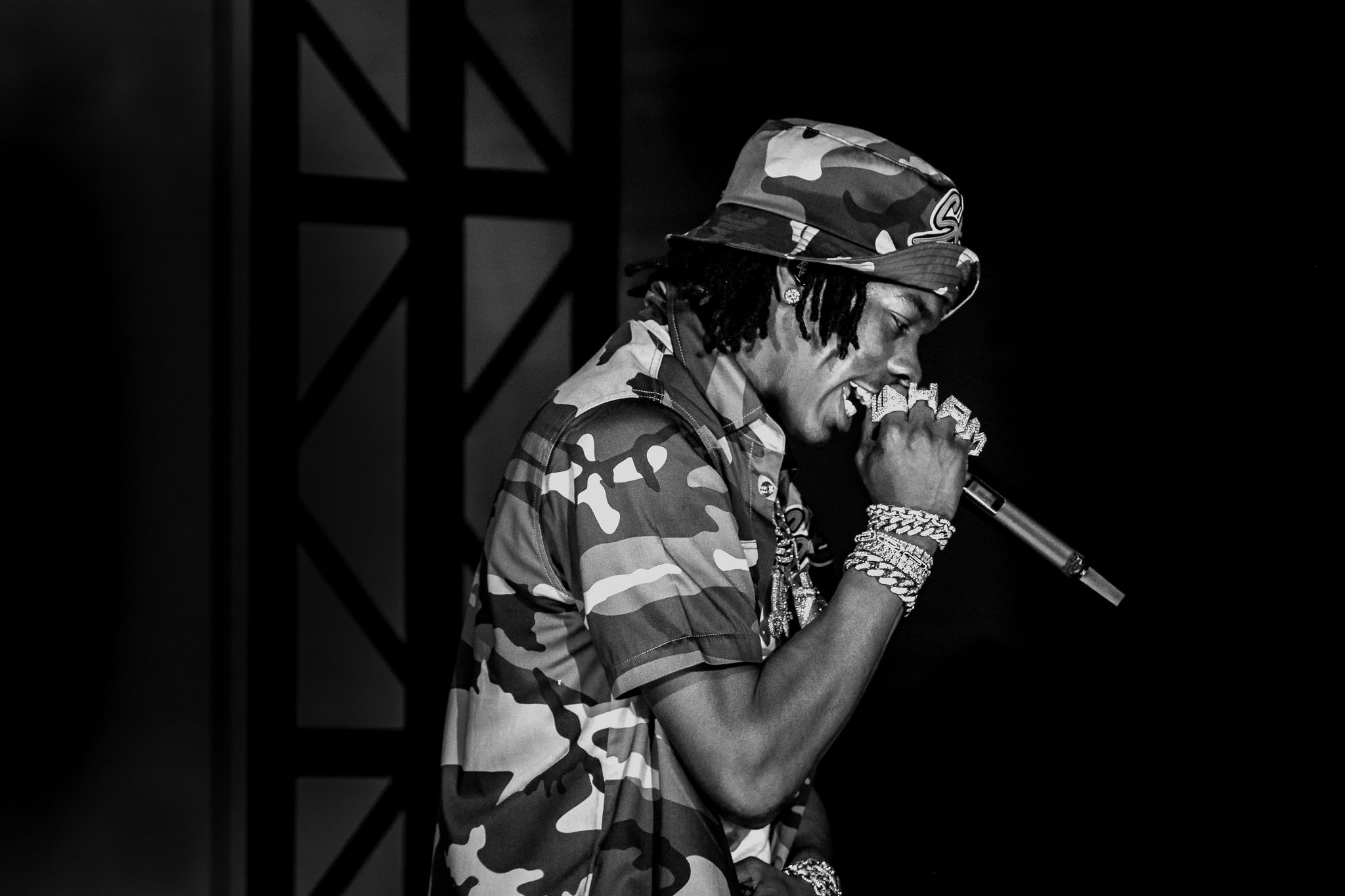 Black and white photo of performer in camo outfit singing into microphone on stage.