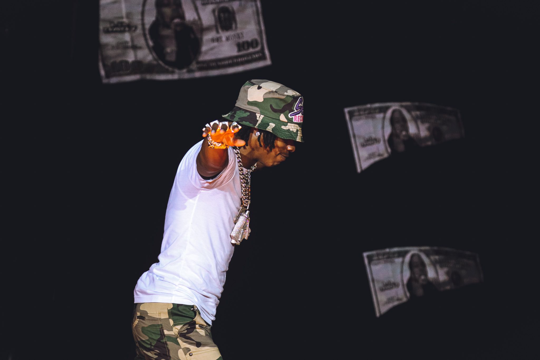 Performer in white shirt and camo pants leans back dramatically on stage with money effects.