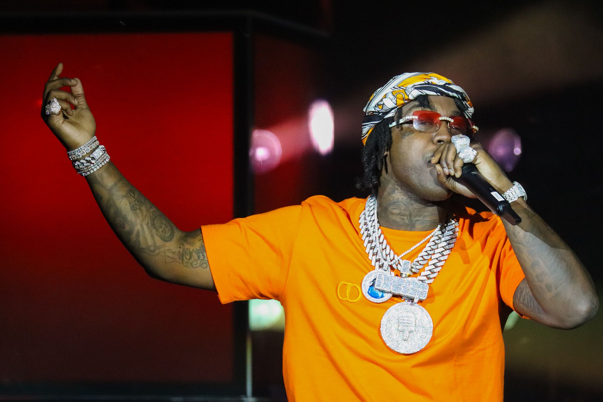 Concert performer in orange shirt and chains gestures while holding microphone on stage.