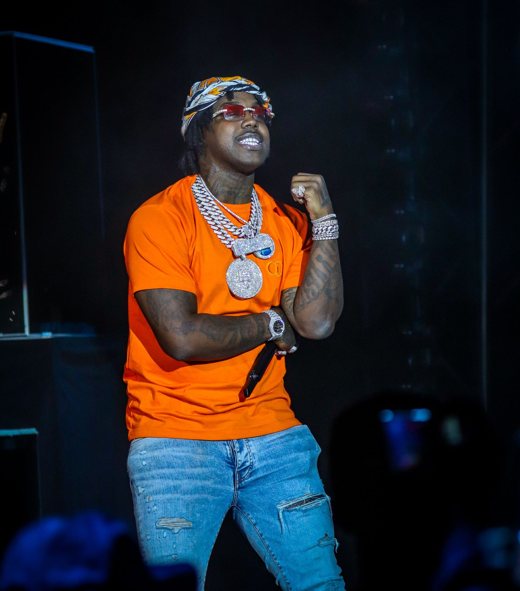 Performer in bright orange shirt and jewelry energetically performs on darkened stage.