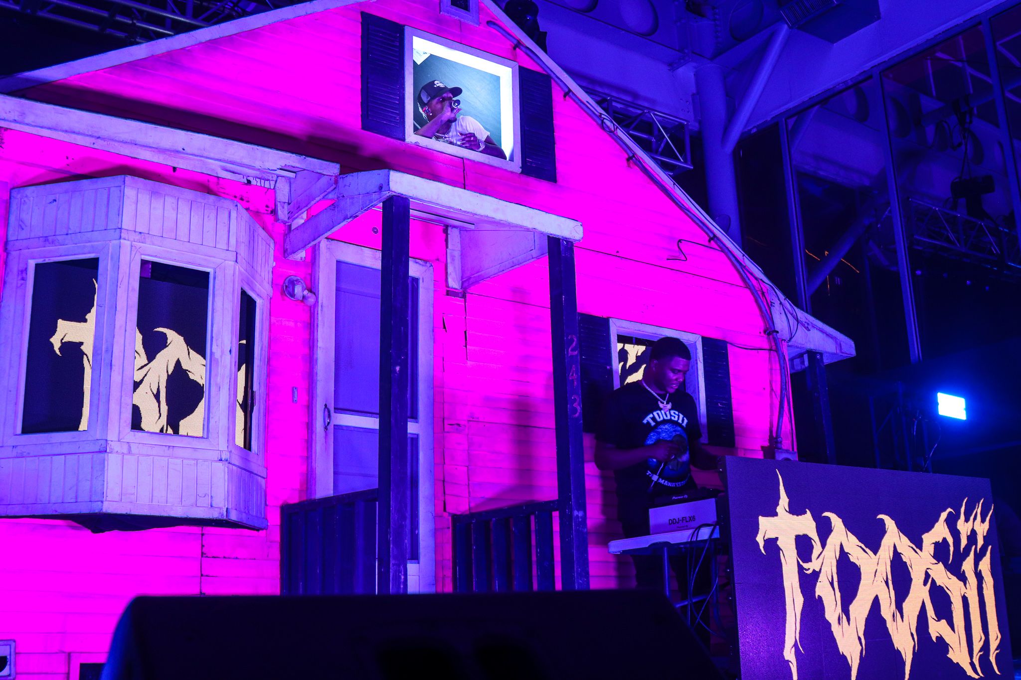 Pink-lit stage set featuring small house facade with performers visible in windows.
